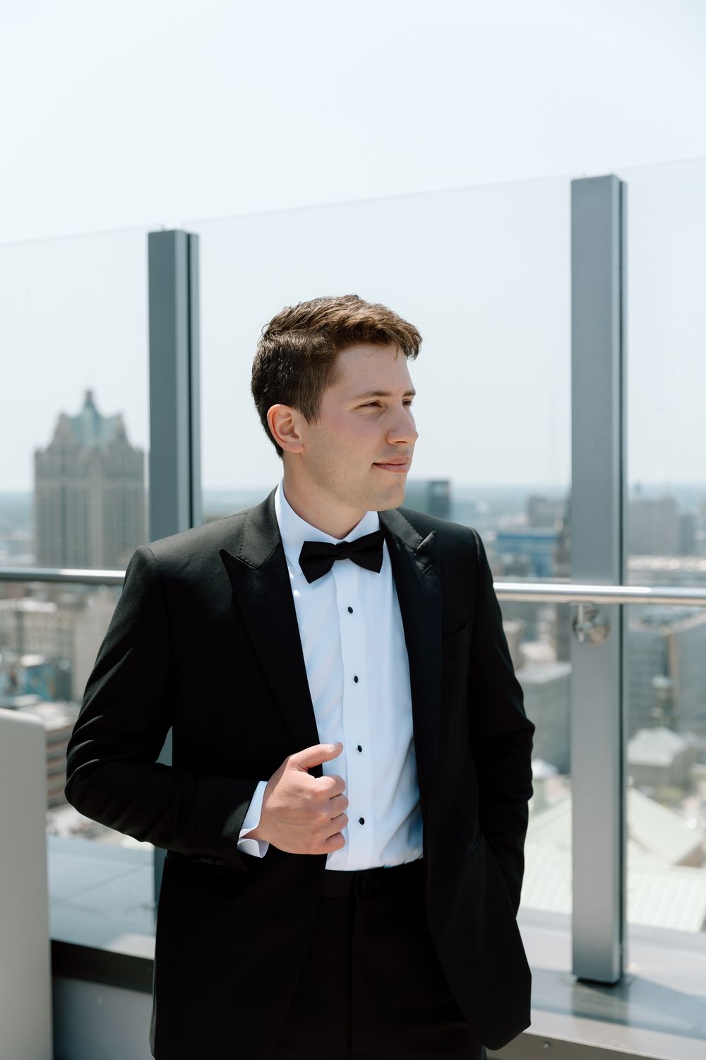 Elegant groom in black tuxedo and bow tie stands on rooftop balcony with city skyline behind him.