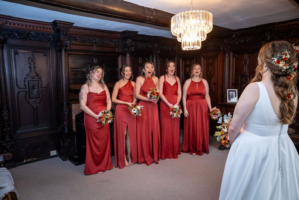 Five bridesmaids in red gowns holding colorful bouquets react with joy seeing the bride in her wedding dress.
