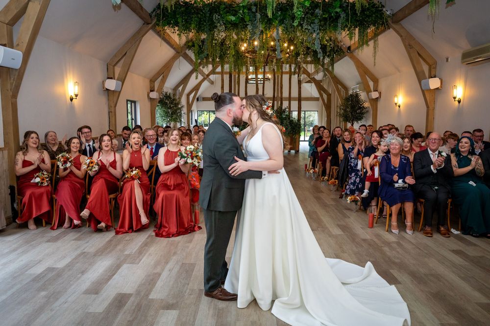 Newlyweds share first kiss in stunning barn venue with red bridesmaid dresses and floral ceiling.
