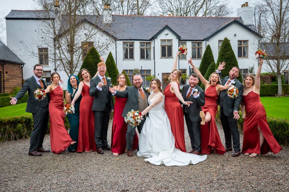 Wedding party poses joyfully in red and green attire outside white country wedding venue.