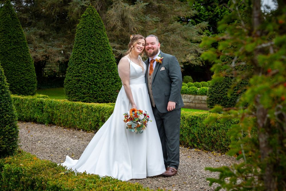 Bride and groom smiling together in a formal garden with manicured hedges and topiary trees on their wedding day.