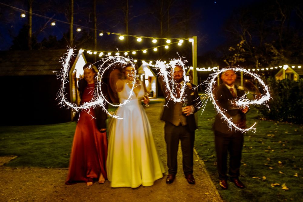 Wedding guests writing LOVE with sparklers at night under string lights during an outdoor evening celebration.