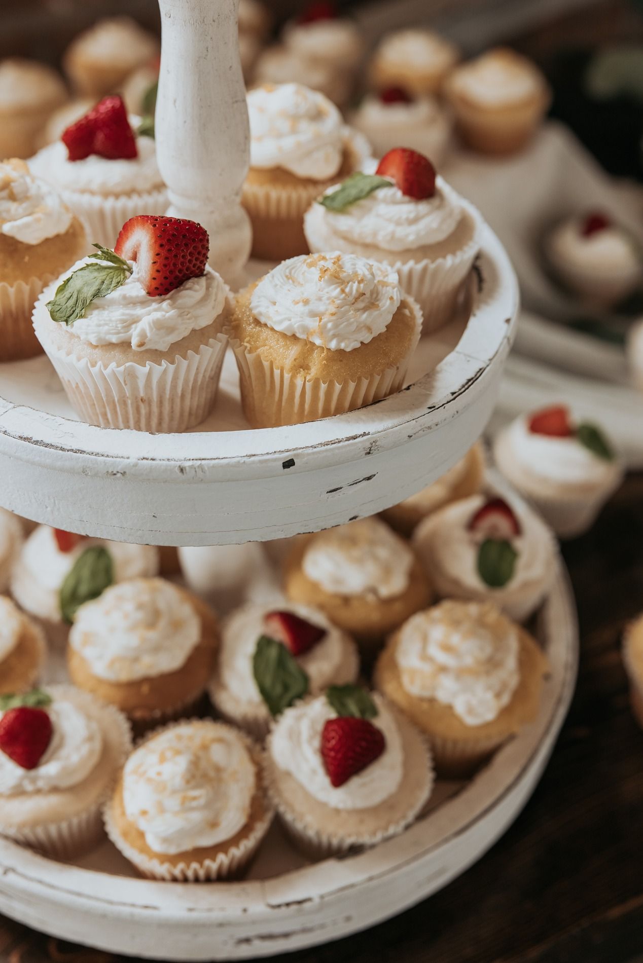 White tiered dessert stand displaying delicate cupcakes topped with whipped cream and fresh strawberries.