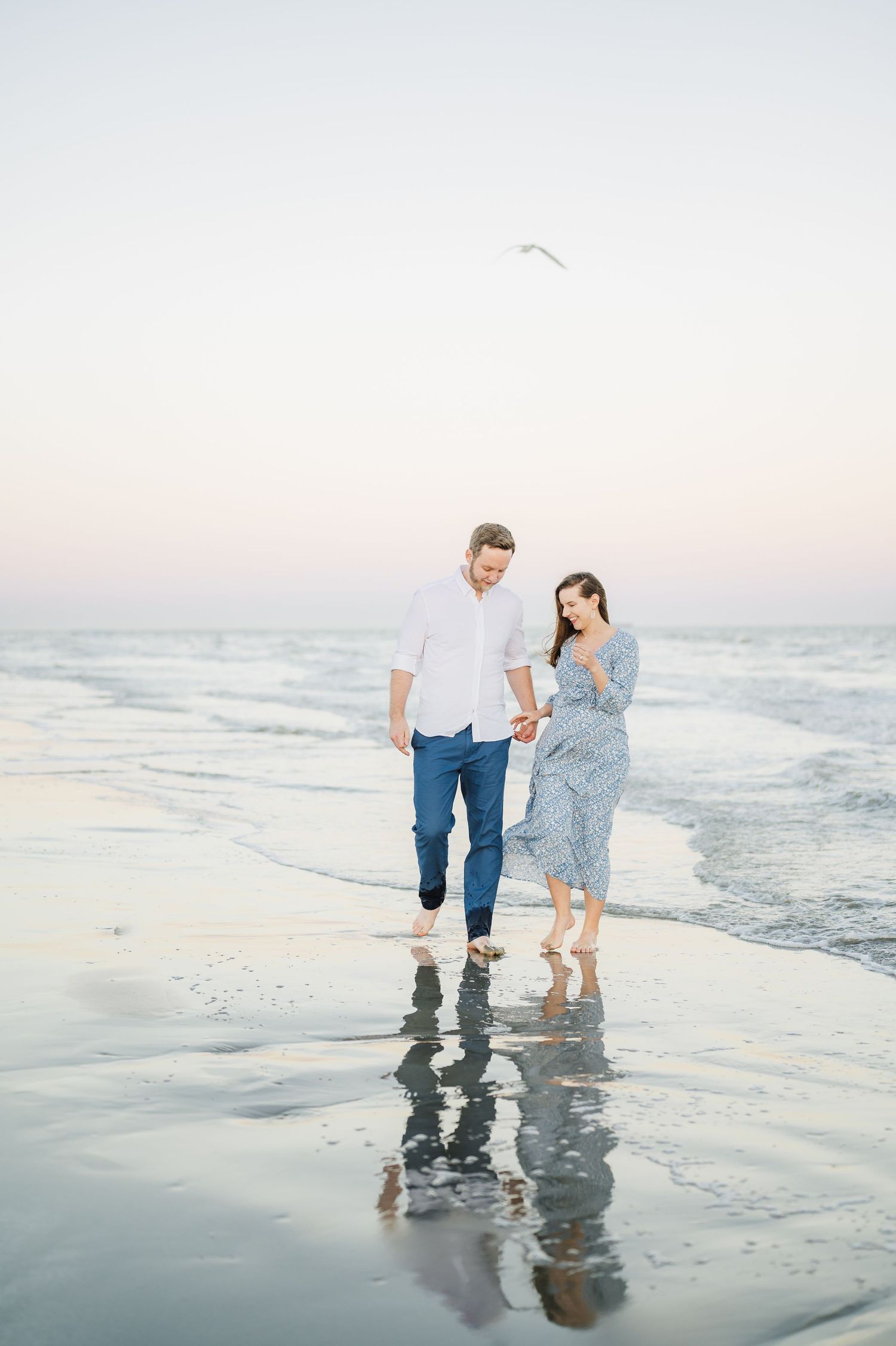 A couple walks along a misty beach shoreline at sunset, their reflections mirrored in the wet sand.