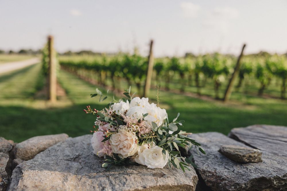 A romantic floral arrangement with blush roses rests on a stone wall in a vineyard setting.