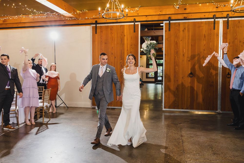 Wedding couple makes their grand entrance into rustic barn reception venue with wooden doors and string lights.