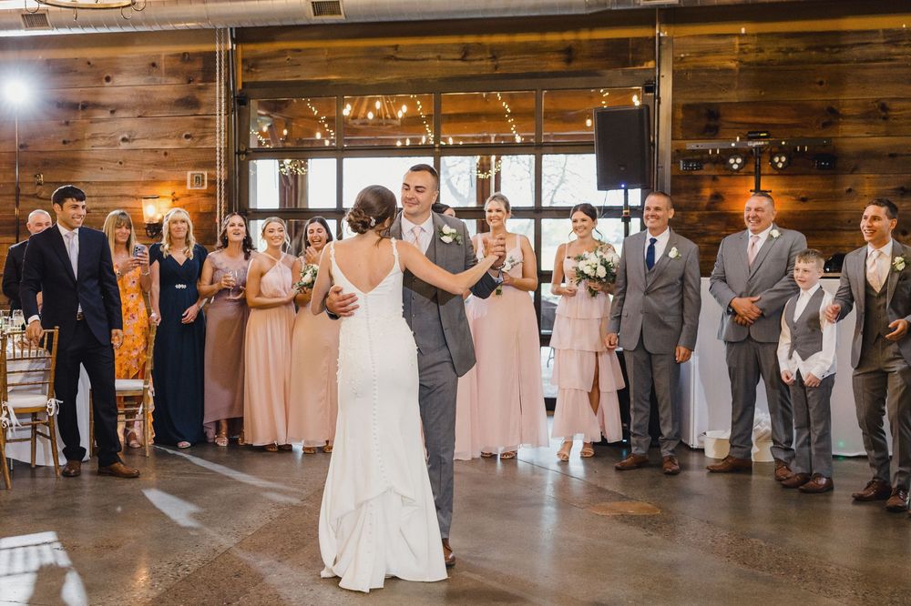 Couple shares their first dance in a rustic venue with string lights and wedding party looking on.