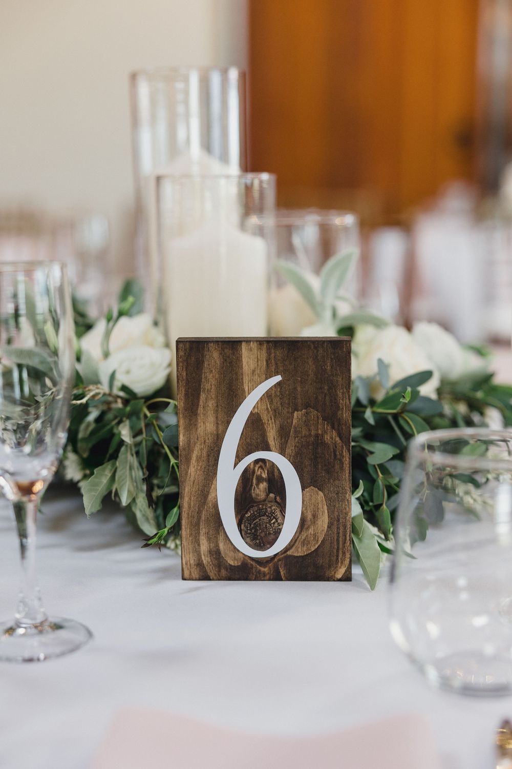 Rustic wooden table numbers and greenery garland decorate an elegant white wedding reception tablescape.