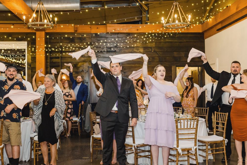 Wedding guests dance and celebrate on a rustic wooden dance floor under twinkling string lights at a barn reception.