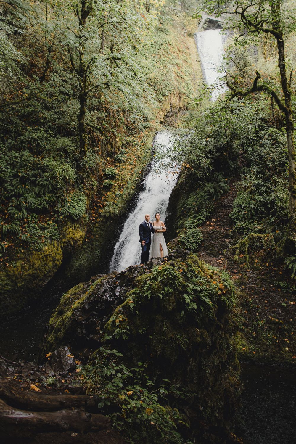 Two figures stand together on mossy rocks in front of a cascading waterfall surrounded by lush green forest.