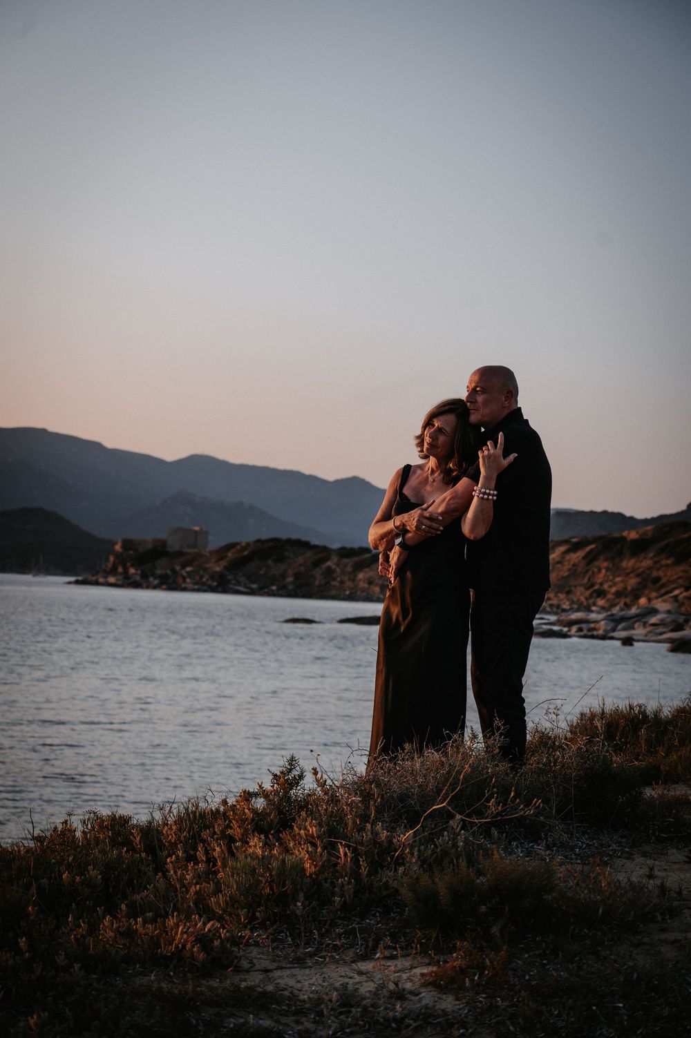 Paar im Sonnenuntergang am Meer mit Bergen im Hintergrund, warmes Abendlicht und ruhige Atmosphäre.