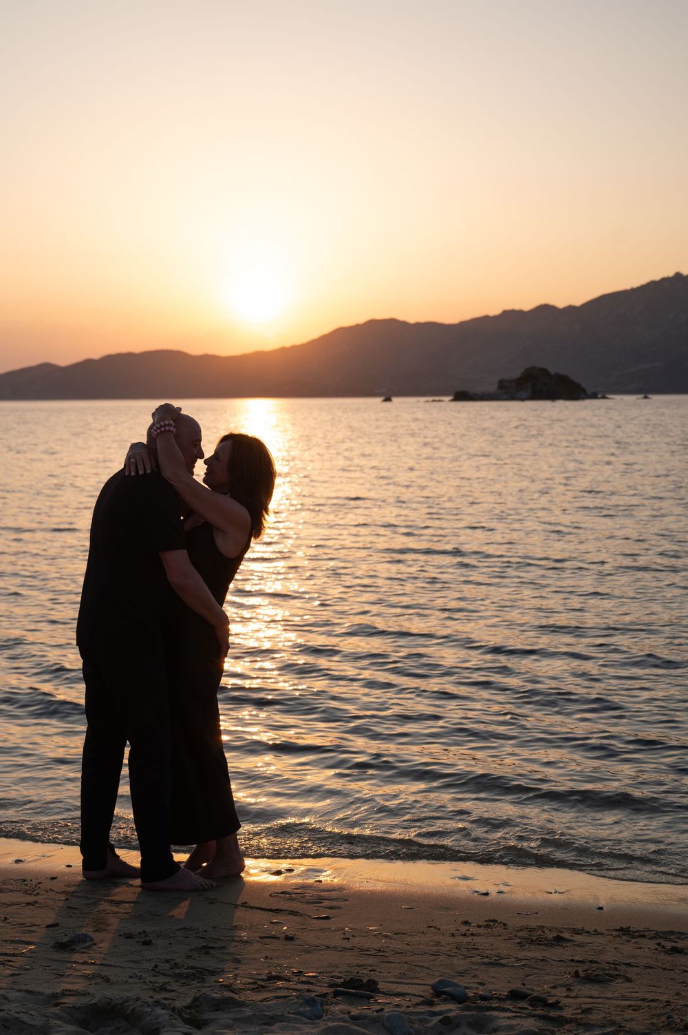 Pärchen im Gegenlicht des Sonnenuntergangs am Meer mit sanften Bergen im Hintergrund.