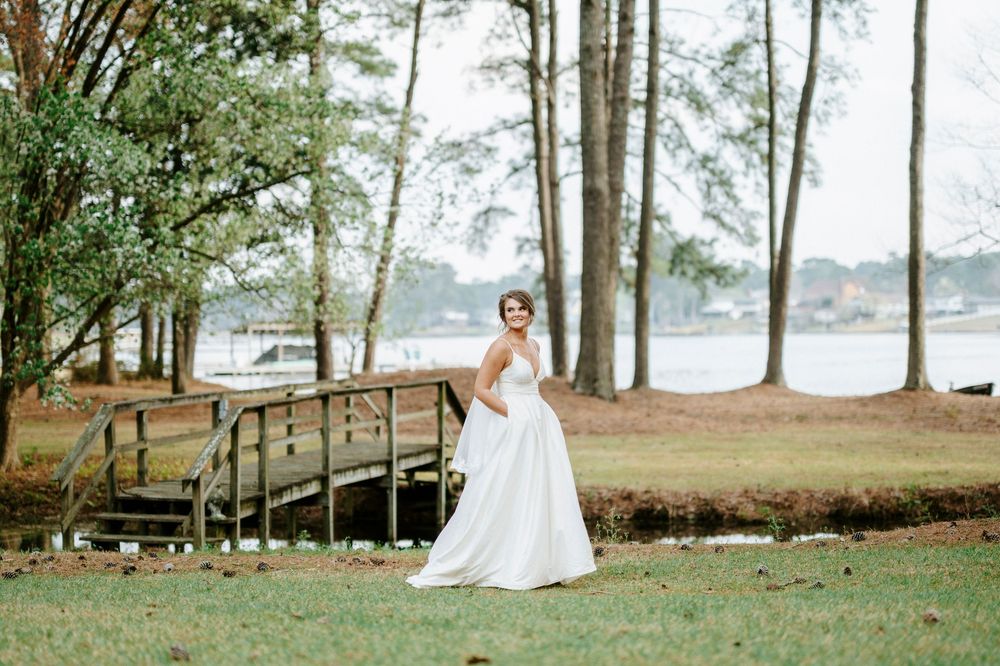 A bride in a flowing white gown poses gracefully in a wooded lakeside setting during a romantic outdoor photo session.