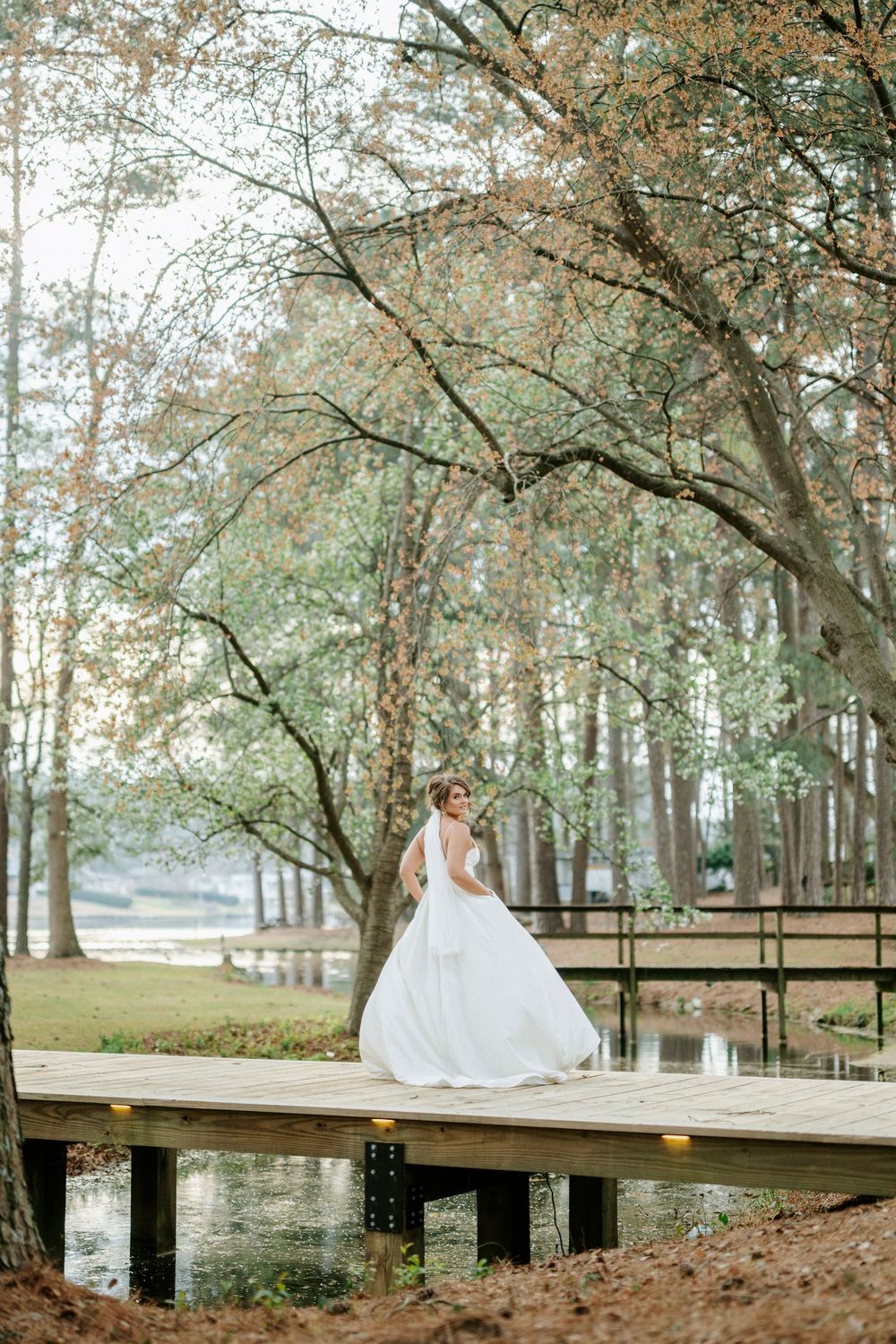 A figure in a flowing white wedding gown stands gracefully on a wooden dock surrounded by autumn trees near a lake.