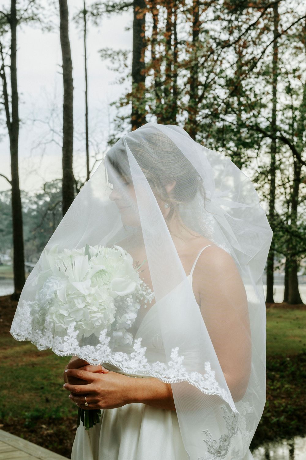 Delicate lace veil with floral embroidery details flows gracefully in a natural outdoor setting with pine trees and water.