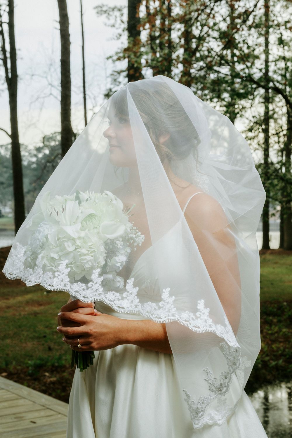 Delicate lace trim detail on flowing white wedding dress veil captured in outdoor woodland setting near water.