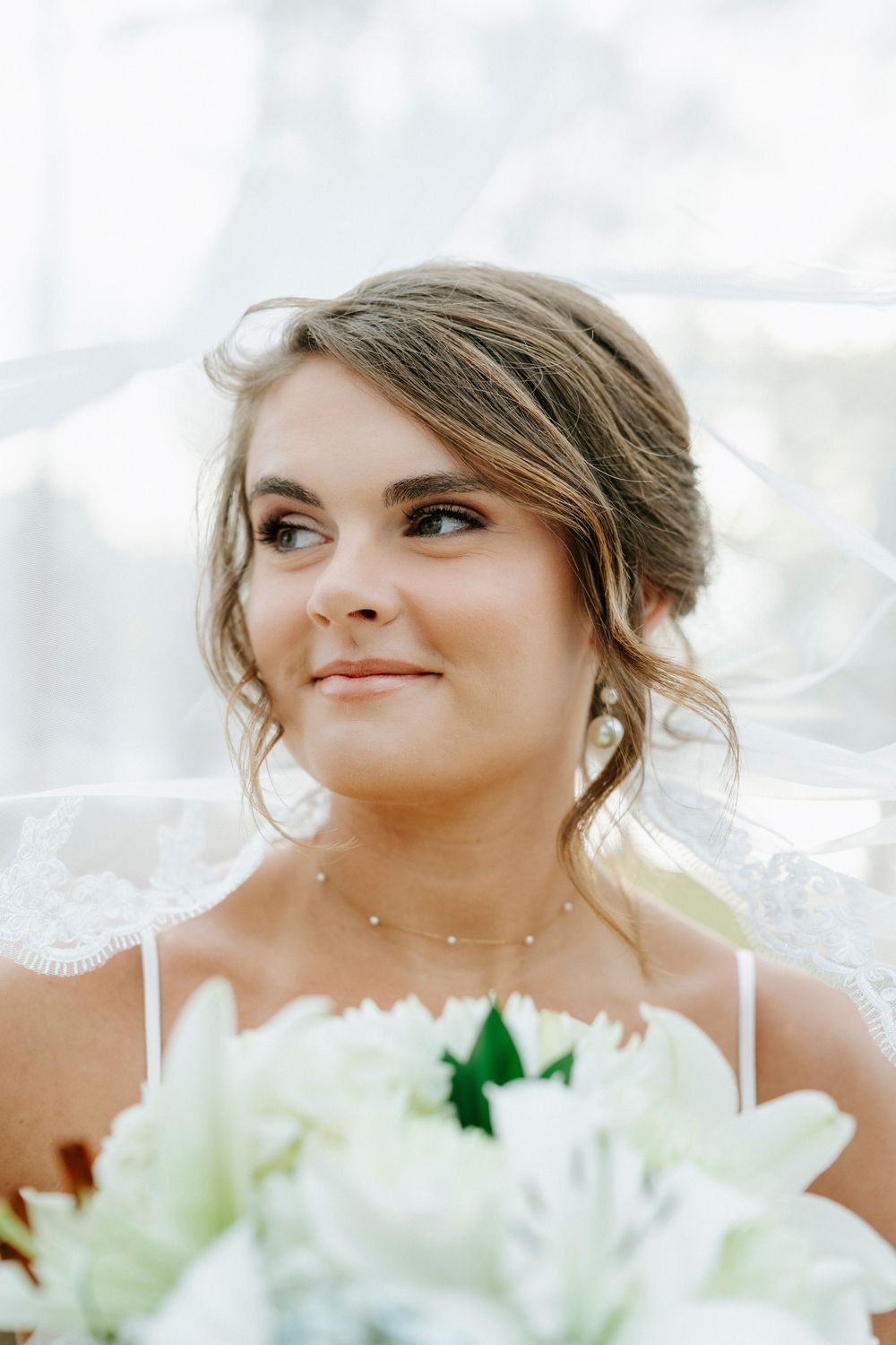 A bride in white dress smiles while holding a bouquet of white flowers against a bright background.