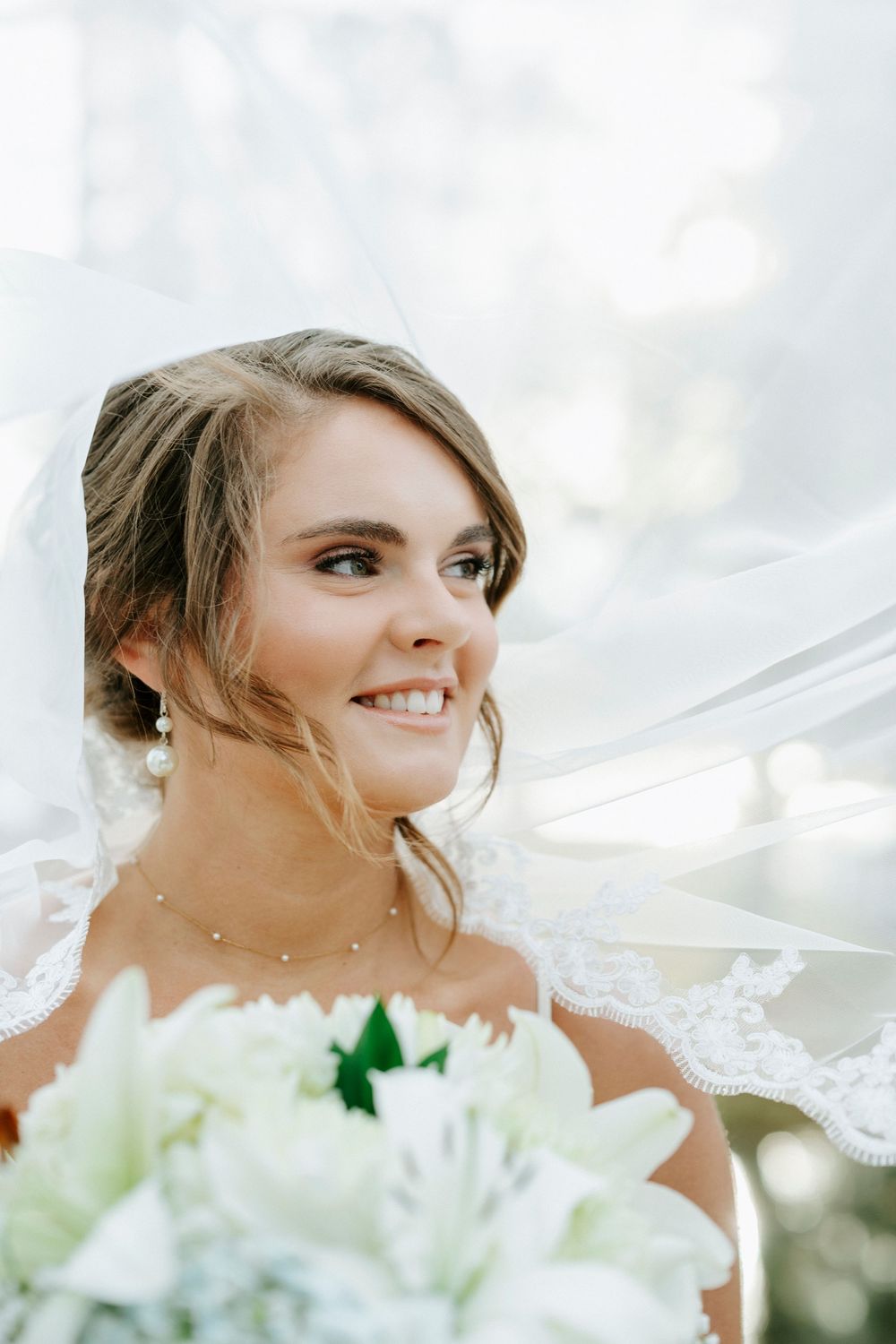A smiling bride holds a white bouquet while her veil flows in the breeze on her wedding day.