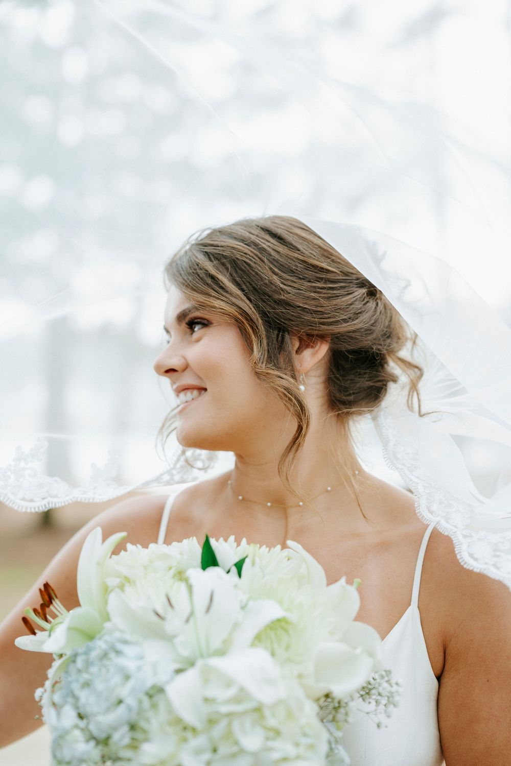 A bride in a white dress holds a bouquet of white flowers while smiling and looking to the side.