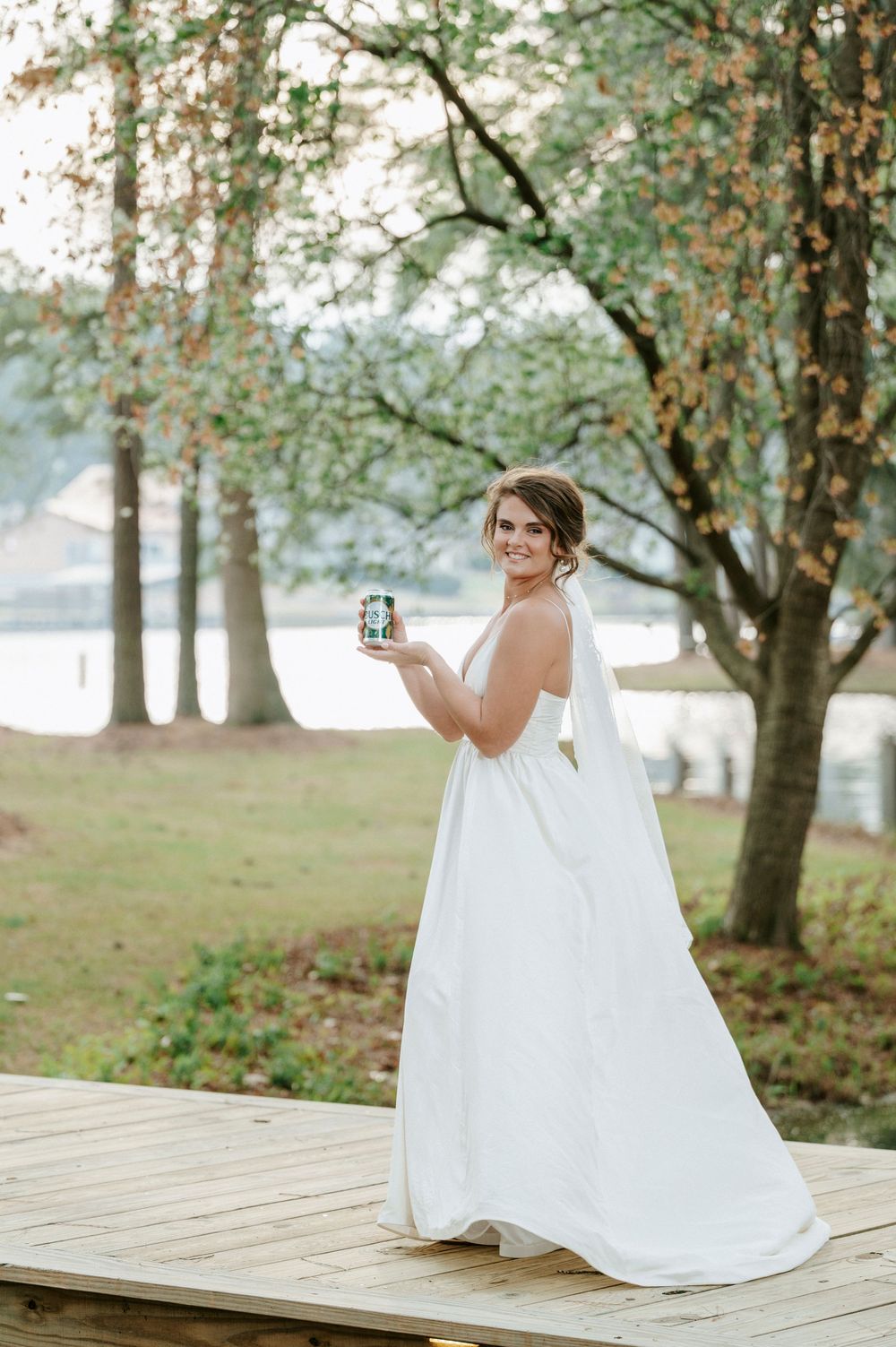 A bride in a flowing white gown sits on a wooden dock by a serene lake surrounded by autumn trees.