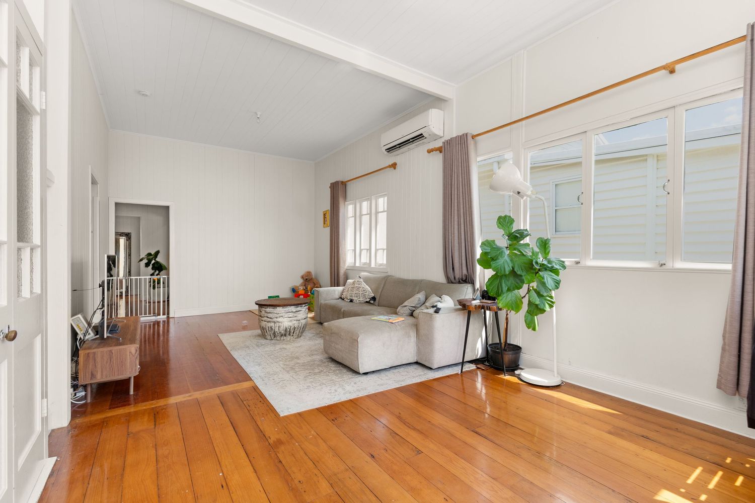 Bright living room with polished timber floors, grey sofa, split system air conditioner, and large windows.