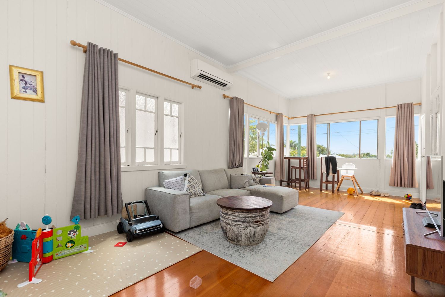 Family living room with grey sofa, polished timber floors, split system AC, and childrens toys visible.