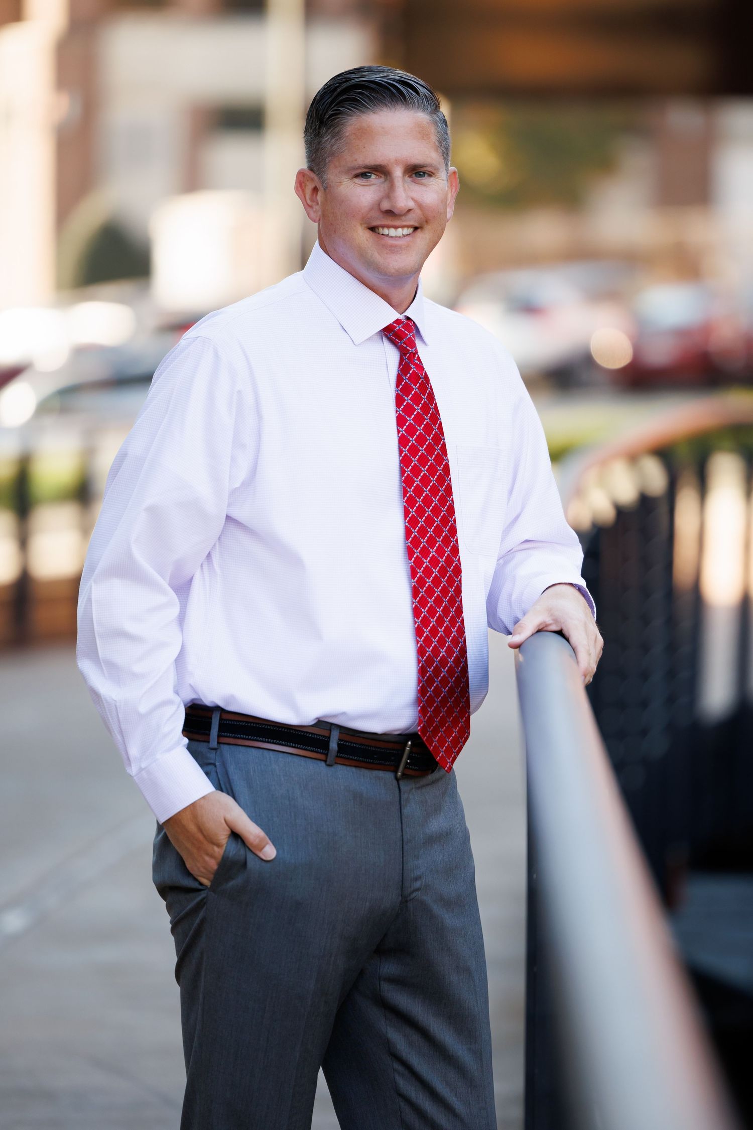 Professional portrait of someone in business attire with a white dress shirt, red patterned tie and gray slacks leaning on railing.