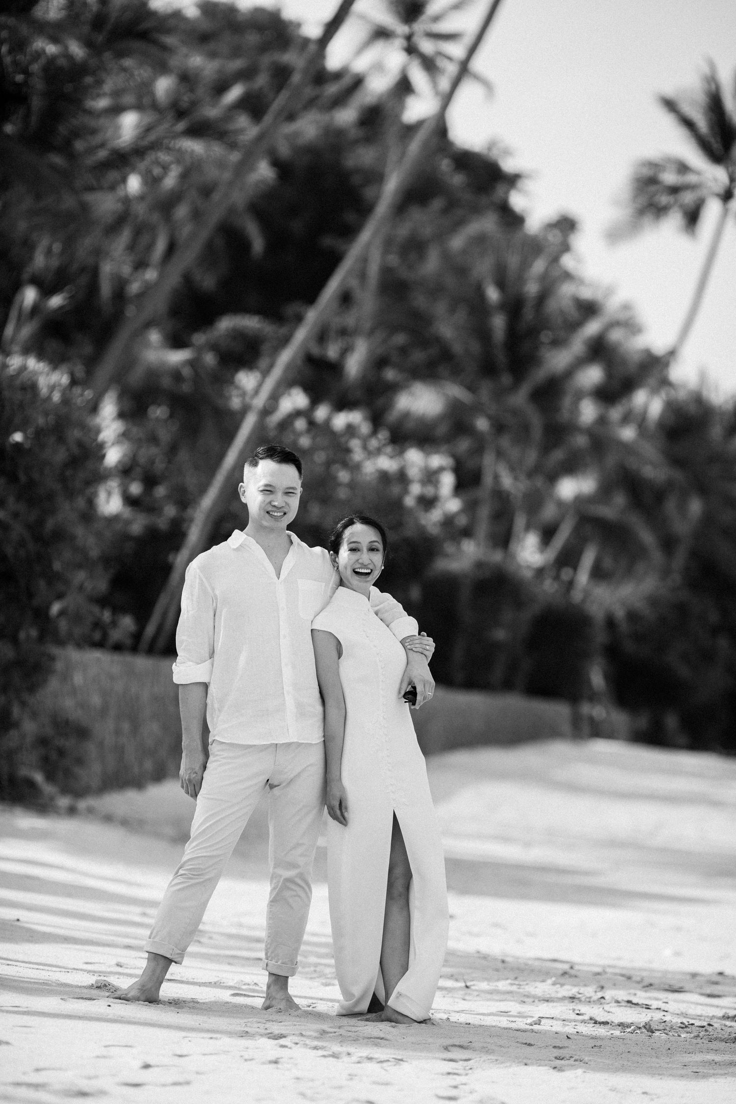 Bride in a white cheongsam posing with groom on a tropical beach in Koh Samui, captured in a timeless black-and-white wedding portrait.