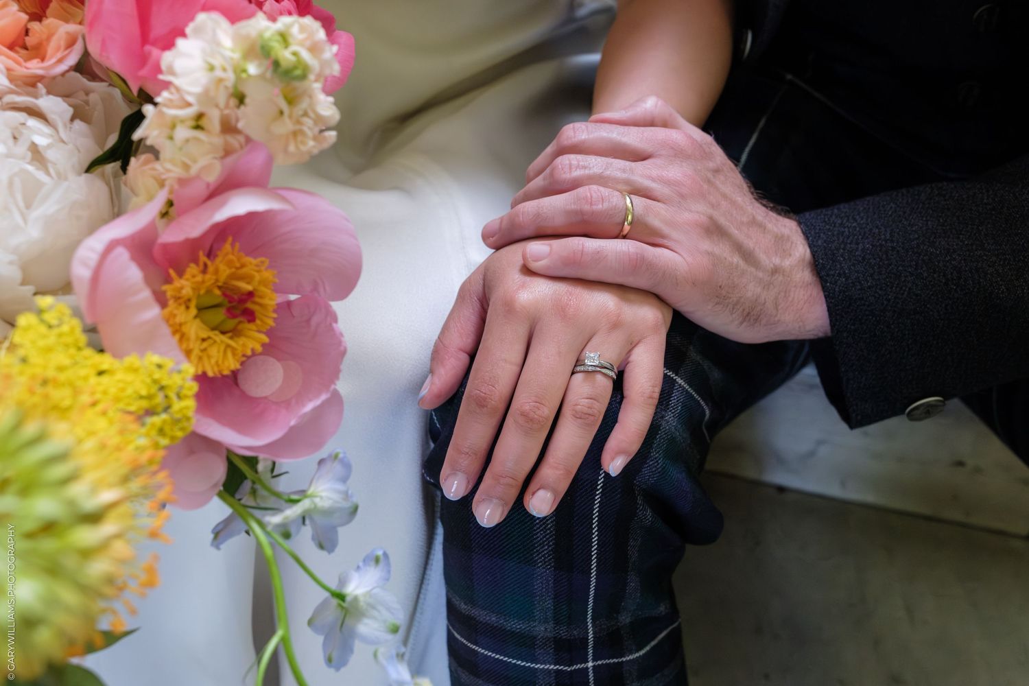 Couple's hands with wedding rings resting on a plaid kilt beside a colorful bouquet of pink peonies and yellow flowers.
