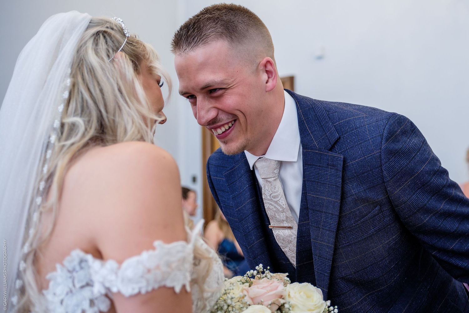Groom in blue suit laughing joyfully with bride in lace wedding dress during wedding ceremony.