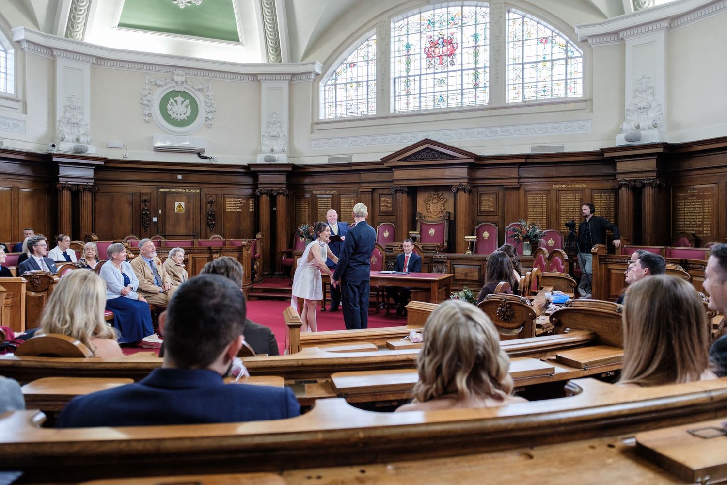 A crowded courtroom with people seated in wooden benches, a judge's bench, and large arched windows.