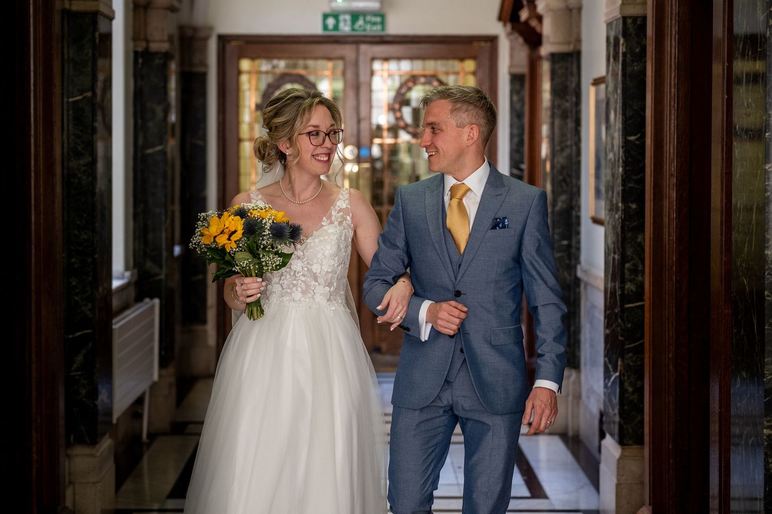A bride and groom walking down a hallway, the bride holding a bouquet of sunflowers.