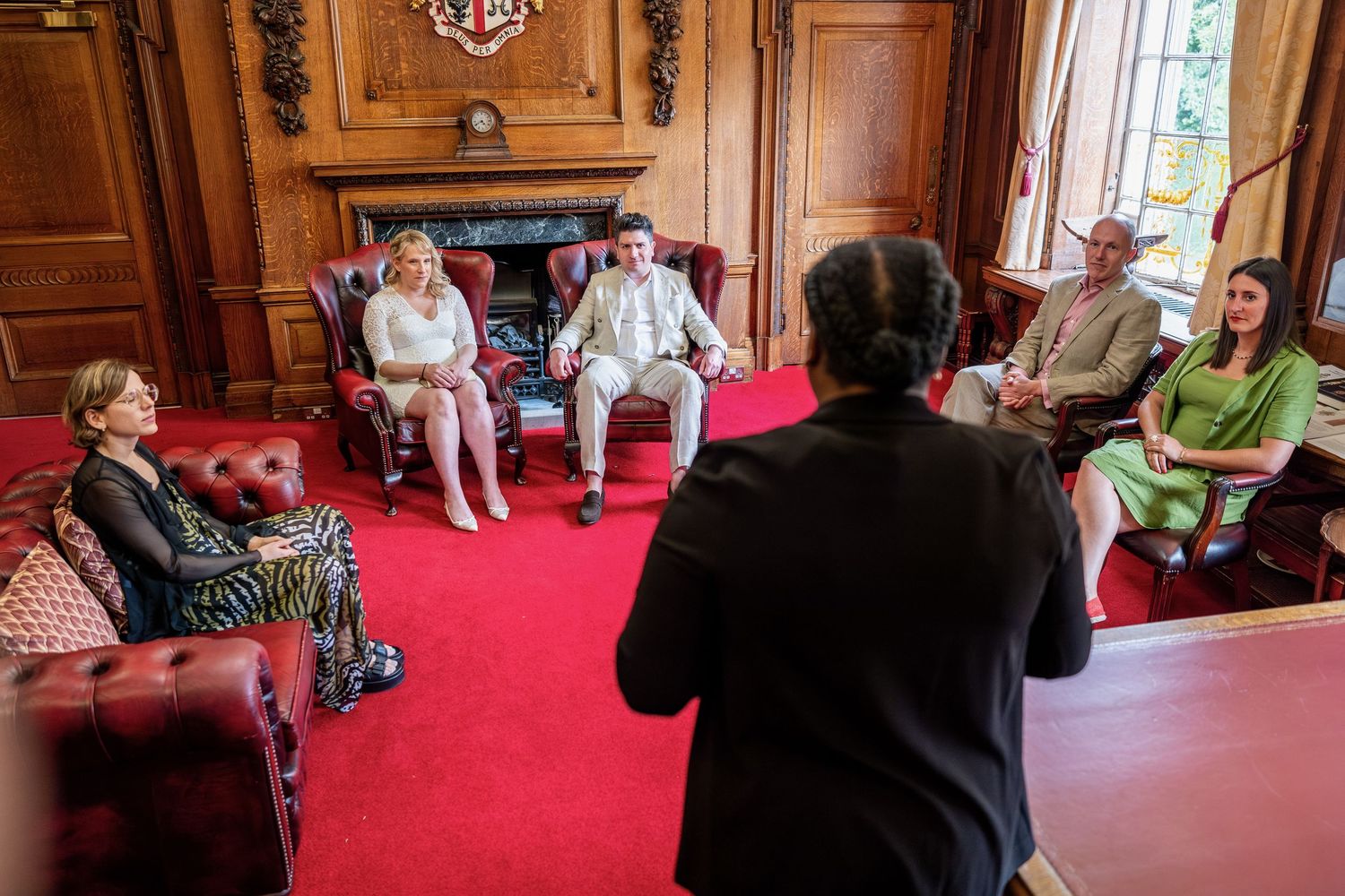 A group of people sitting and conversing in a formal, ornate interior setting with red carpeting and wooden furnishings.