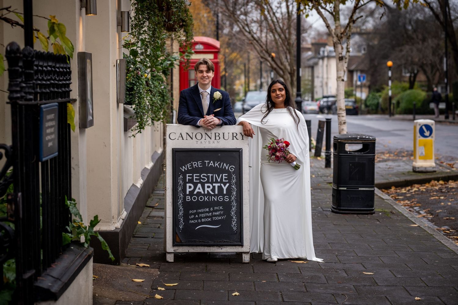 A bride and groom stand in front of a sign advertising a party in a city street.