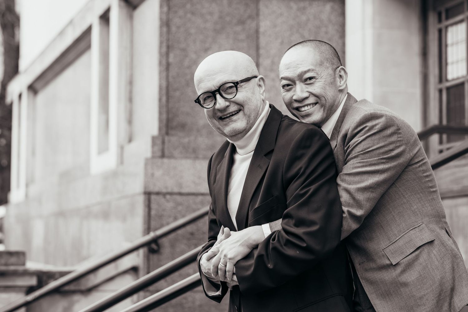 Two older men in suits and glasses stand together, smiling and embracing on a city street.