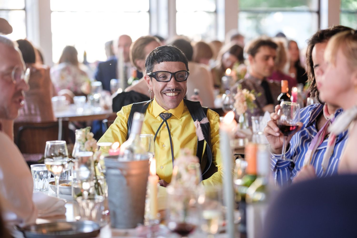 A man wearing glasses and a yellow shirt is standing at a bar surrounded by other people.