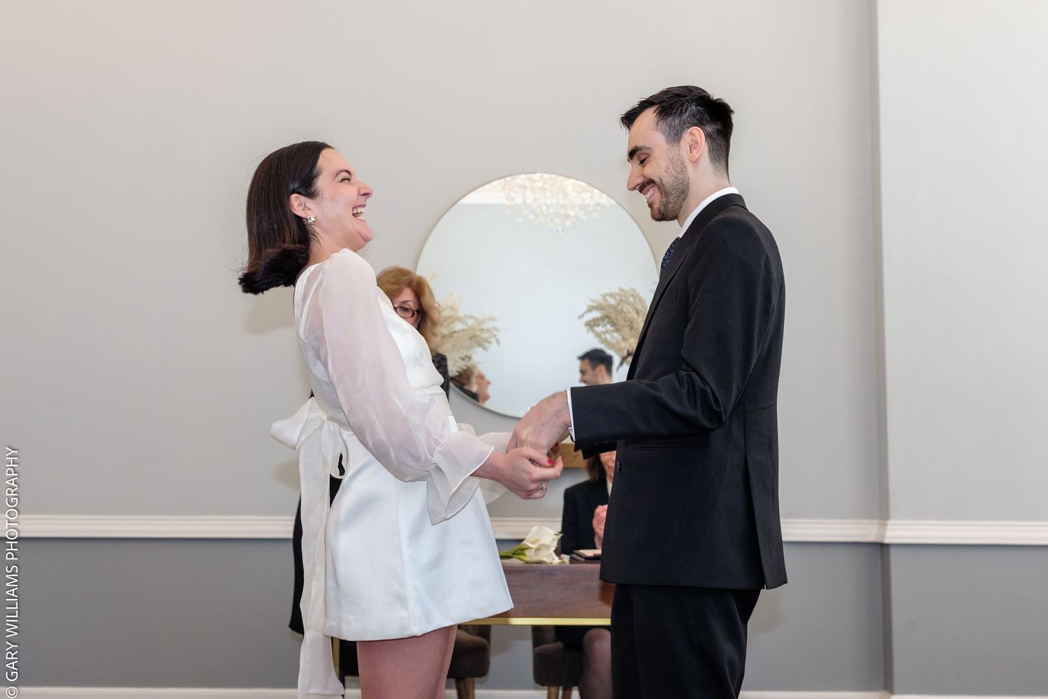Couple holding hands and smiling during an intimate indoor wedding ceremony, bride in white dress, groom in black suit.