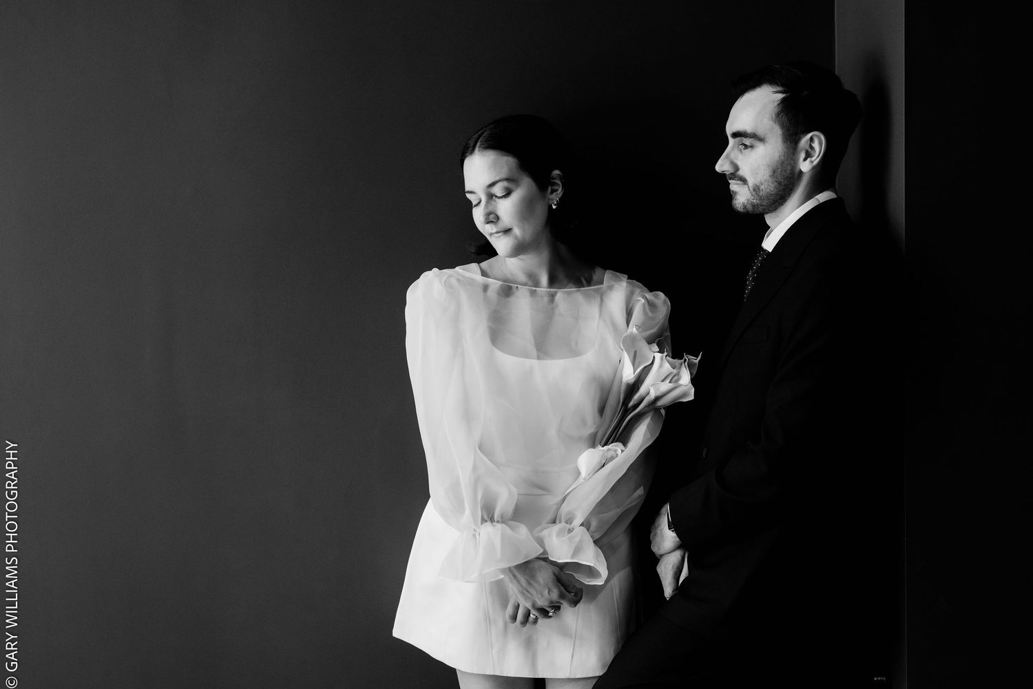 Black and white wedding photo of bride holding bouquet in white dress beside groom in tuxedo against dark wall.