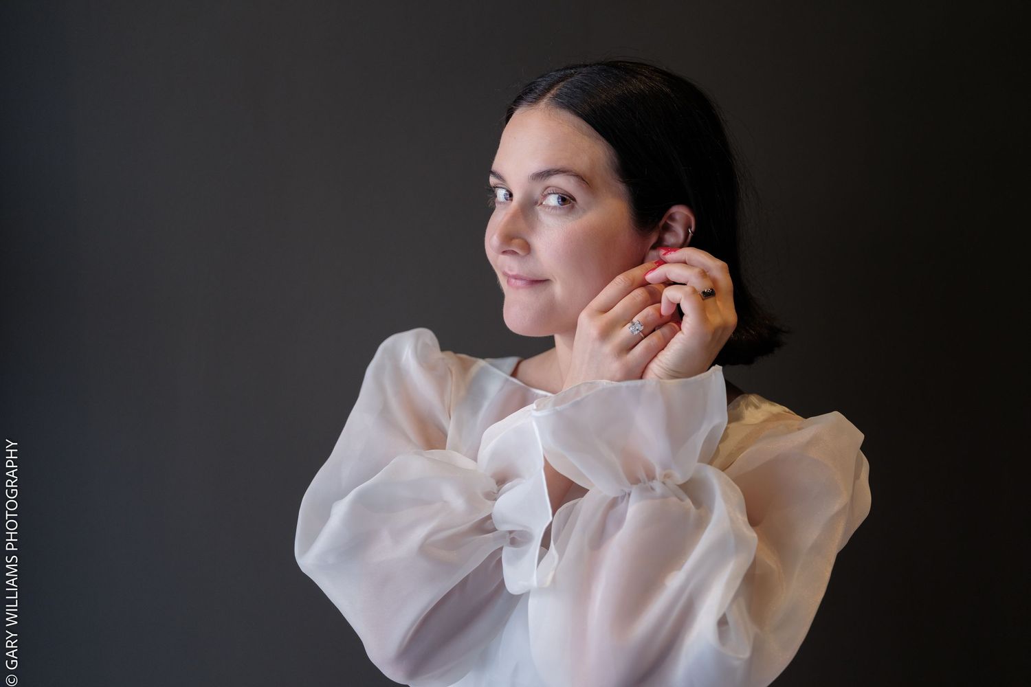 Woman in white ruffled blouse posing elegantly against dark background, touching her ear with ring-adorned hand.