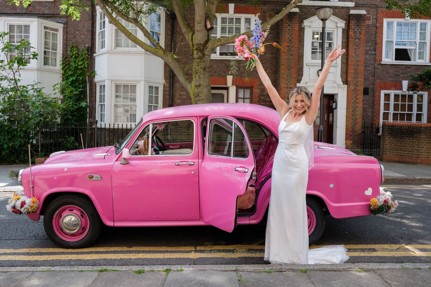 A woman in a white wedding dress poses next to a vibrant pink vintage car on a city street.