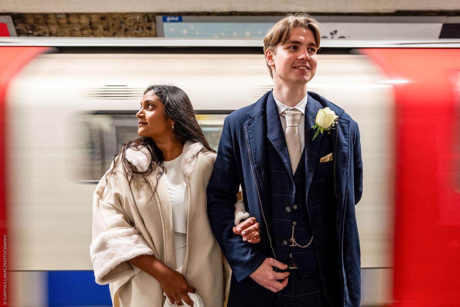 A young couple stands together in front of a red train, the woman wearing a light-colored coat and the man in a suit.