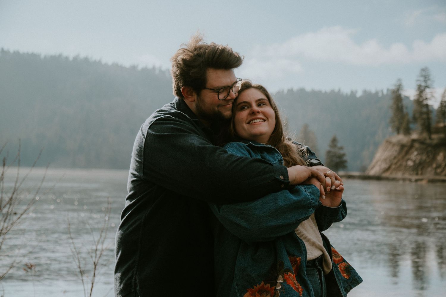 A couple embracing each other while standing near a body of water surrounded by trees.