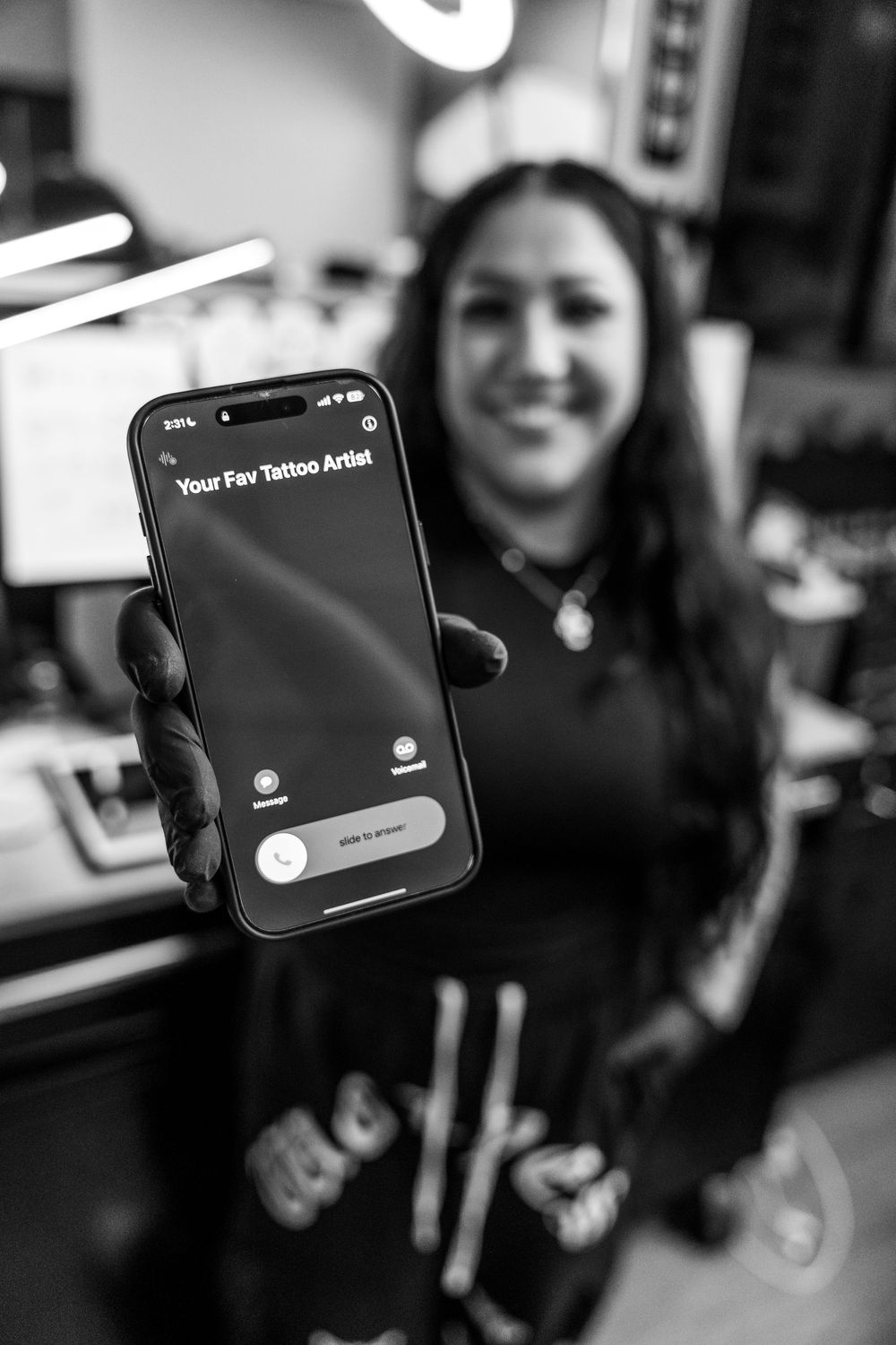 Smiling tattoo artist holding phone displaying Your Fav Tattoo Artist screen in black and white. Jacksonville, NC