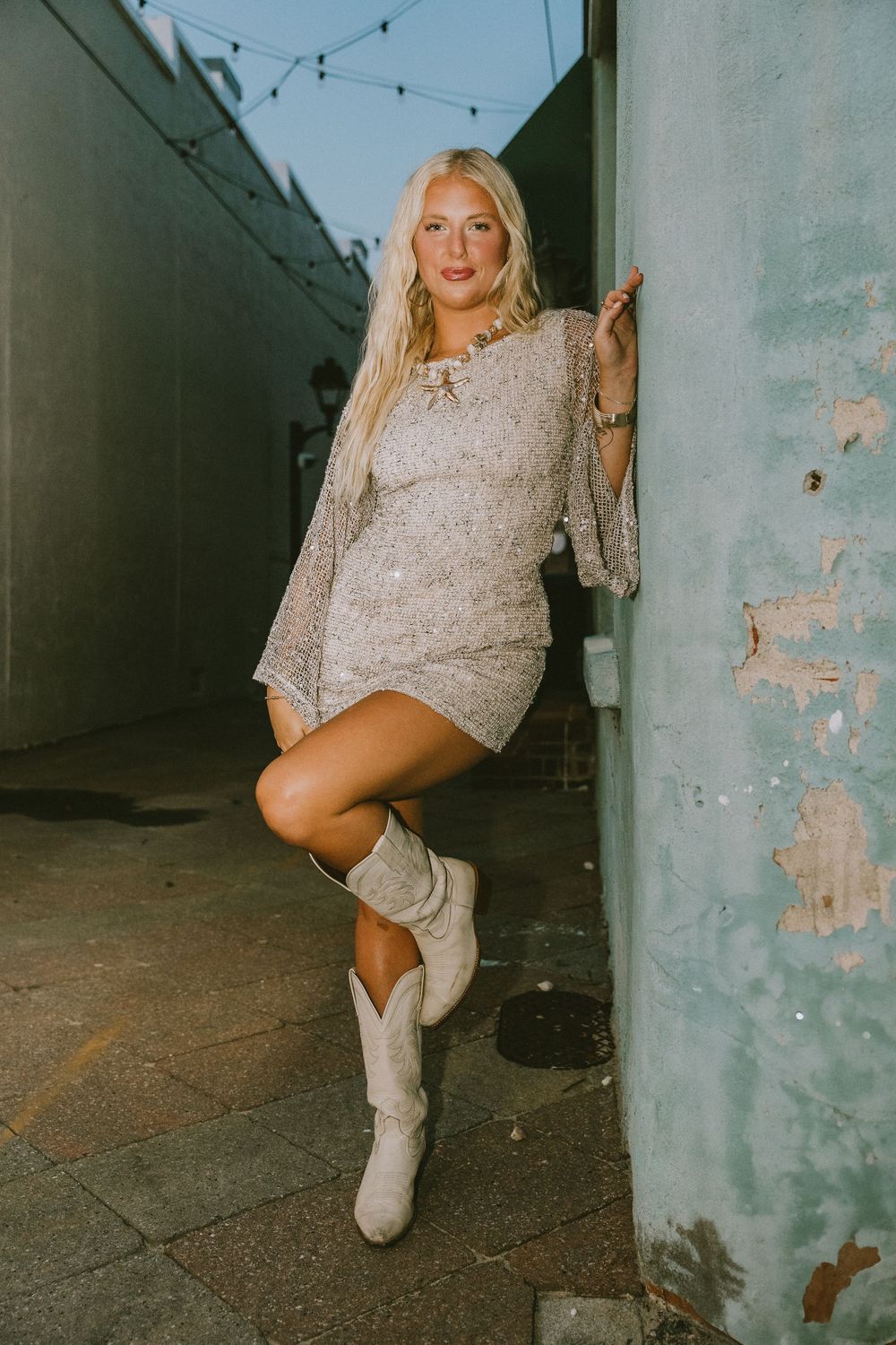 Blonde woman in sequin mini dress and cream cowboy boots posing against weathered wall in alley. Greenville, NC