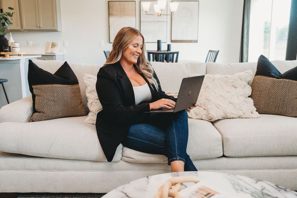 Smiling blonde woman working on laptop while sitting on modern white sofa in stylish living room. Jacksonville, NC
