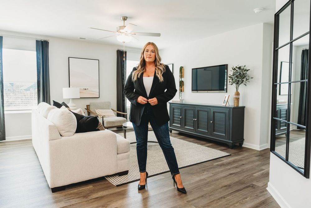 Confident blonde woman in black blazer and jeans standing in modern living room with hardwood floors. Real estate agent branding photos.