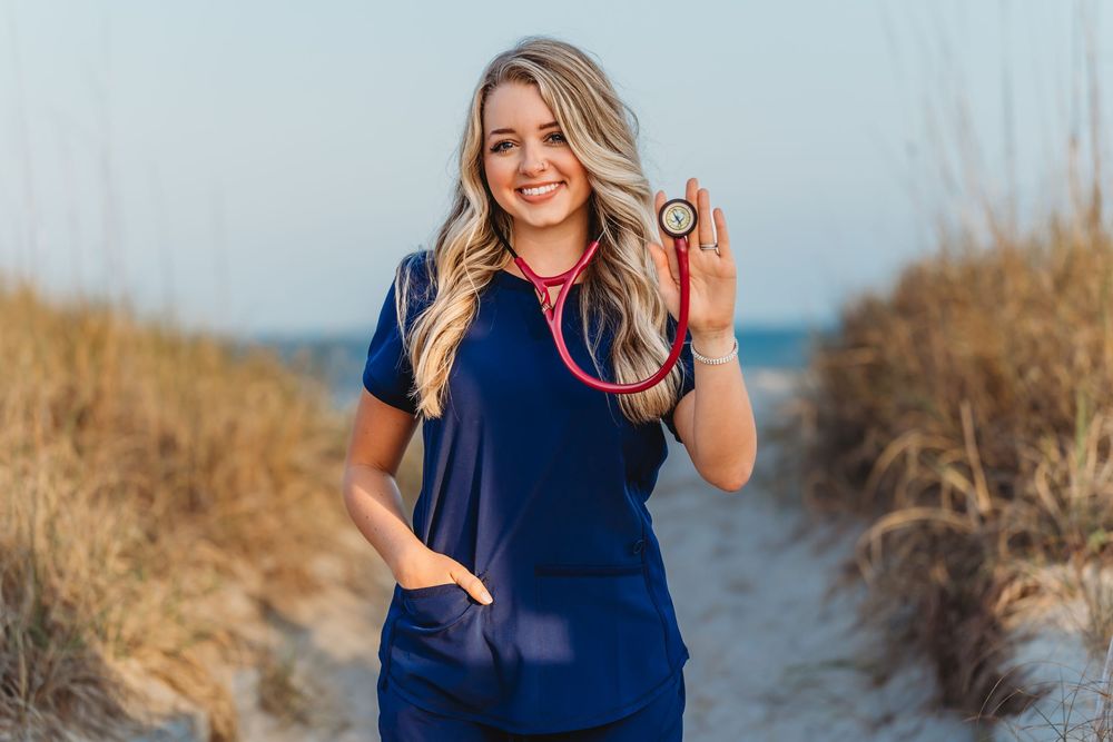 Smiling blonde woman in navy scrubs holding red stethoscope on sandy beach path. Camp Lejeune, NC