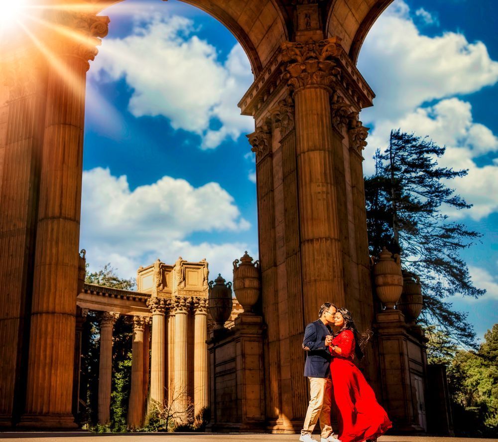 Couple in formal attire embracing outdoors near trees and a wooden bench on a gravel path.