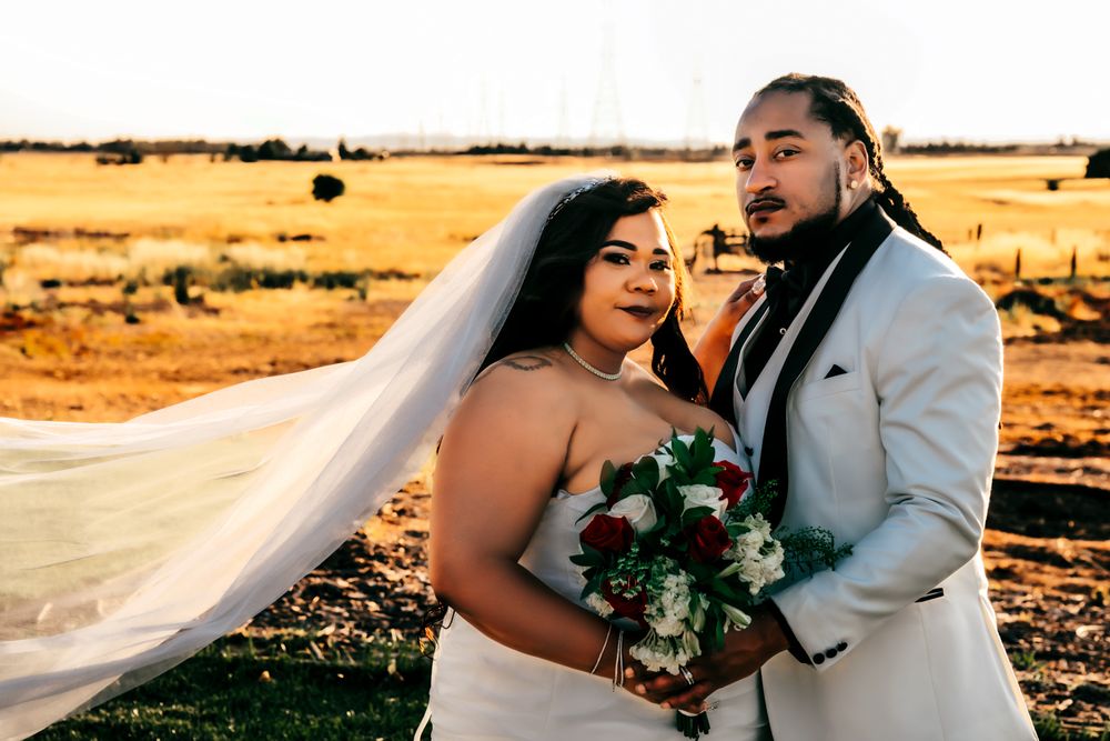 Bride in white gown and veil holds bouquet beside groom in white suit at golden hour outdoor wedding.