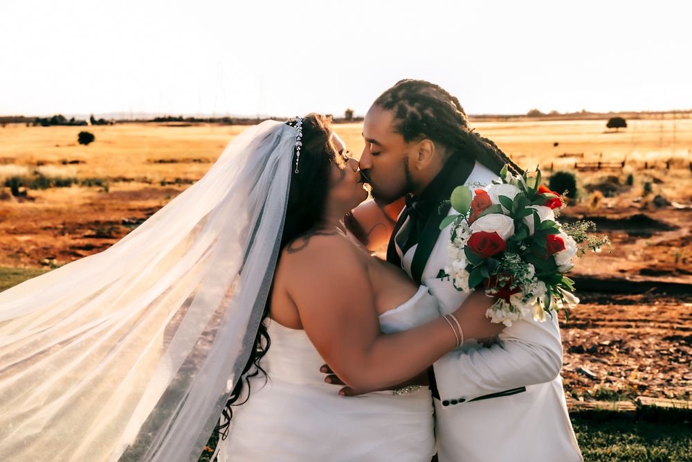 Bride and groom sharing a romantic kiss outdoors at golden hour, with a flowing veil and red bouquet.